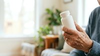 Senior man with brown skin holding white cosmetic bottle in home interior with natural light and plants visible in background.