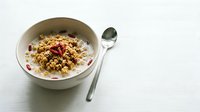 Overhead view of granola cereal with yogurt and berries in white bowl with spoon on white surface, breakfast food styling.