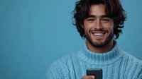 Young Hispanic man with wavy dark hair wearing knit sweater smiles while holding smartphone against blue background, expressing joy and confidence in casual studio portrait.