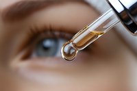 Close-up of glass dropper with golden liquid drop against blurred eye background. Macro shot capturing skincare serum or essential oil application in cosmetics.