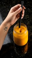 Hand holding reusable metal straw in glass jar of fresh orange juice on dark surface. Caucasian person with geometric tattoo on wrist enjoying sustainable beverage lifestyle.