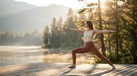 Young woman practicing yoga warrior pose on wooden dock at mountain lake during golden hour sunrise with peaceful forest landscape background.