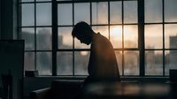 Silhouette of contemplative man sitting by large industrial window during golden hour sunset creating moody atmospheric scene for mental health concepts.