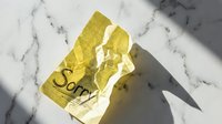 Crumpled yellow sticky note with the word Sorry written on white marble surface with natural shadows, symbolizing regret and apology.