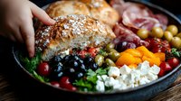 Mediterranean bowl with fresh bread, olives, feta cheese, tomatoes, and mixed vegetables served on rustic wooden table, close up view of colorful ingredients.