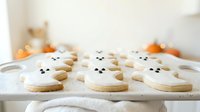 Cute ghost-shaped sugar cookies with almond eyes arranged on white cloth with autumn pumpkins in soft-focus background, perfect for fall baking, holiday treats, and seasonal dessert content.