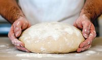 Baker kneading fresh dough on wooden surface, hands covered in flour, close-up view showing texture and preparation process for homemade bread making.