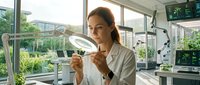 Young female scientist examining laboratory glassware in modern research facility with natural lighting and green outdoor view.