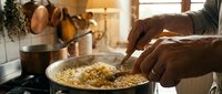 Hands stirring cooked pasta in a pot on stovetop in warm kitchen setting with cookware and natural light for cooking and food preparation.