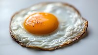 Close-up of sunny side up fried egg with crispy edges and bright orange yolk on light background, showing texture and detail in morning breakfast cooking.