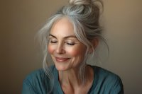 Mature Caucasian woman with silver gray hair in messy bun smiling peacefully with eyes closed wearing teal shirt against neutral beige background for wellness content.