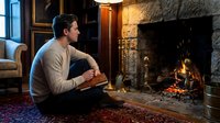 Young Caucasian man in beige sweater reading book by stone fireplace in cozy traditional living room with warm lighting and classic decor.