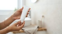 Woman with medium skin tone holding white cosmetic tube in bathroom setting, displaying skincare or beauty product for packaging mockup and promotional use.