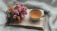 Cup of herbal tea placed on open book pages with dried pink and orange flowers arrangement on soft gray textile background, creating cozy reading atmosphere.