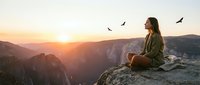 Young woman hiker sitting on mountain cliff edge at sunrise watching birds fly over dramatic canyon landscape during peaceful morning meditation moment.