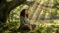 Young woman meditating peacefully under ancient tree with sunbeams filtering through branches, creating serene forest atmosphere for wellness and mindfulness.