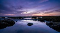 Tranquil lake reflects dramatic purple sunset sky with scattered rocks and marsh grass creating peaceful natural landscape for relaxation and meditation themes.