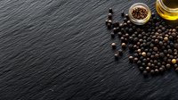 Black peppercorns scattered on dark slate stone surface with glass jar of spice and olive oil bottle, copy space on left side for text placement.