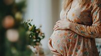 Expecting mother in floral dress holds belly while standing near window with soft bokeh lights. Gentle natural morning light creates peaceful maternal atmosphere.