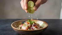 Hands squeezing fresh lime juice over ceviche dish with seafood, vegetables and herbs in wooden bowl for cooking and food preparation.