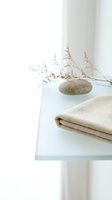 Minimalist spa setup with folded beige towel, smooth stone and dried botanical branches on frosted glass shelf against white wall background for wellness and relaxation design.