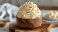 Homemade whipped coconut cream topped with toasted coconut flakes in wooden bowl on rustic cutting board, creamy dairy-free dessert topping.