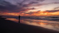 Silhouette of person standing on beach at sunset with dramatic orange and purple sky reflecting on wet sand and ocean waves.
