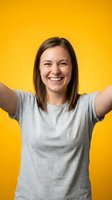 Young woman with shoulder-length brown hair and Caucasian ethnicity wearing gray t-shirt, arms raised celebrating with genuine smile against bright yellow background.