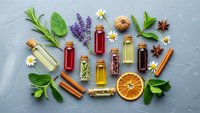 Natural essential oils in glass bottles with fresh herbs, spices and dried orange slice arranged on textured gray background for aromatherapy and wellness.