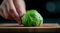 Fresh Brussels sprout on wooden cutting board with hand placing vegetable, dramatic dark background with soft natural lighting and shallow depth of field.