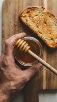 Hand holding jar of honey with wooden dipper above toasted bread slice on rustic wooden board, perfect for food styling, cooking blogs, breakfast recipes, and culinary content.