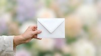 Woman's hand holding white envelope against blurred garden background, suitable for mail, communication, invitation, and greeting card concepts.