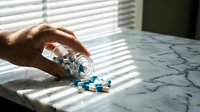 Adult male hand pouring blue and white capsule pills from glass bottle onto marble surface in bright sunlight, suitable for pharmaceutical, healthcare, and medical supplement marketing materials.