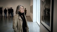 Mature blonde woman in beige coat admiring artwork in modern art gallery museum with visitors in background exploring cultural exhibition space.