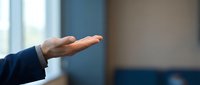 Male hand in dark blue business sleeve extending open palm gesture indoors with blurred office background, conveying offer, presentation, or welcome concept for corporate communications.