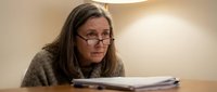 Focused middle-aged Caucasian woman with glasses reviewing documents at desk under warm lamp light, serious expression, indoor office setting.