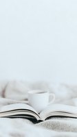 Open book with white coffee cup on soft textured bedding creates cozy reading atmosphere against light background, minimalist lifestyle composition.