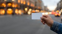 Hand holding blank white card on urban street with blurred evening city lights and historic buildings in background, suitable for business mockups and promotional campaigns.