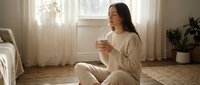 Young Asian woman in cozy beige loungewear sitting on wooden floor holding coffee mug in bright sunlit bedroom with plants creating peaceful morning atmosphere.