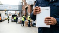 Male construction worker in navy uniform holding blank white clipboard at industrial job site with team and equipment in background, suitable for business, safety, and workplace communications.