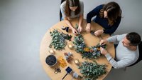 Three women crafting handmade Christmas wreaths with eucalyptus branches and decorative elements on wooden table, holiday DIY workshop activity.