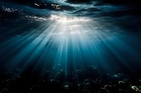 Dramatic sunbeams pierce through ocean surface creating ethereal underwater light rays in deep blue water with rocky seafloor beneath. Perfect for nature themes.