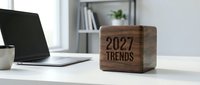 Wooden cube with 2027 trends text on a modern office desk beside a laptop, representing future business planning and forecasting concepts.
