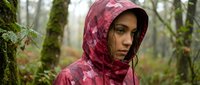Young woman wearing pink camouflage rain jacket with hood in misty forest surrounded by moss-covered trees and vegetation, outdoor adventure and nature exploration.