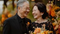 Senior Asian couple sharing joyful moment at autumn wedding celebration, wearing elegant black attire with orange floral decorations and natural outdoor setting.