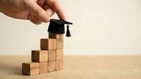 Hand placing a miniature graduation cap on top of stacked wooden blocks forming stairs, symbolizing steps to academic success.