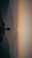 Silhouetted figure standing on rocky cliff edge overlooking misty valley at golden hour sunrise or sunset, atmospheric landscape with warm orange and golden light breaking through fog.