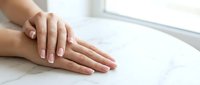 Young woman with natural manicure displaying healthy nails and smooth skin on white marble surface near window for beauty and wellness content.