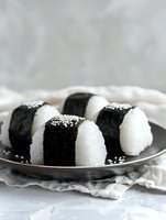 Fresh onigiri rice balls wrapped in crisp nori seaweed, garnished with white sesame seeds on dark ceramic plate against light background. Japanese cuisine.