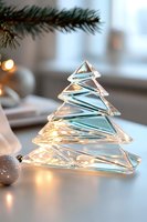 Crystal glass Christmas tree decoration with warm glowing lights and silver ornaments creates elegant holiday atmosphere against soft window light background.
